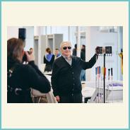 Организатор фестиваля Александр Петрович Митьков стоит в выставочном зале рядом с демонстрационным стендом. В руках он держит стойку с тростями различного цвета. Рядом фотограф делает снимок.